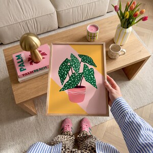 May include: A framed art print of a potted plant with green leaves and white spots, set against a pink and yellow geometric background. The print is held by a person wearing a blue and white striped shirt and pink trainers.