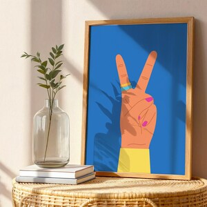 May include: A framed art print featuring a hand making the peace sign against a blue background. The hand is orange with pink fingernails, a gold and blue ring, and a yellow sleeve. The artwork is displayed on a woven table with a vase and books.