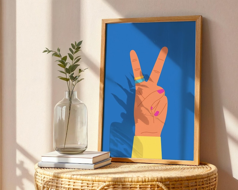 May include: A framed art print featuring a hand making the peace sign against a blue background. The hand is orange with pink fingernails, a gold and blue ring, and a yellow sleeve. The artwork is displayed on a woven table with a vase and books.