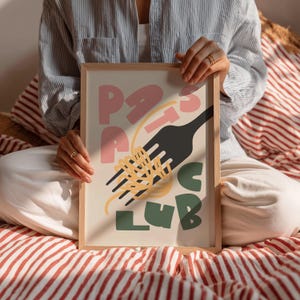 May include: A framed art print with the words "Pasta Club" in pink and green, with a black fork and yellow spaghetti. The print is held by a person sitting on a red and white striped surface.