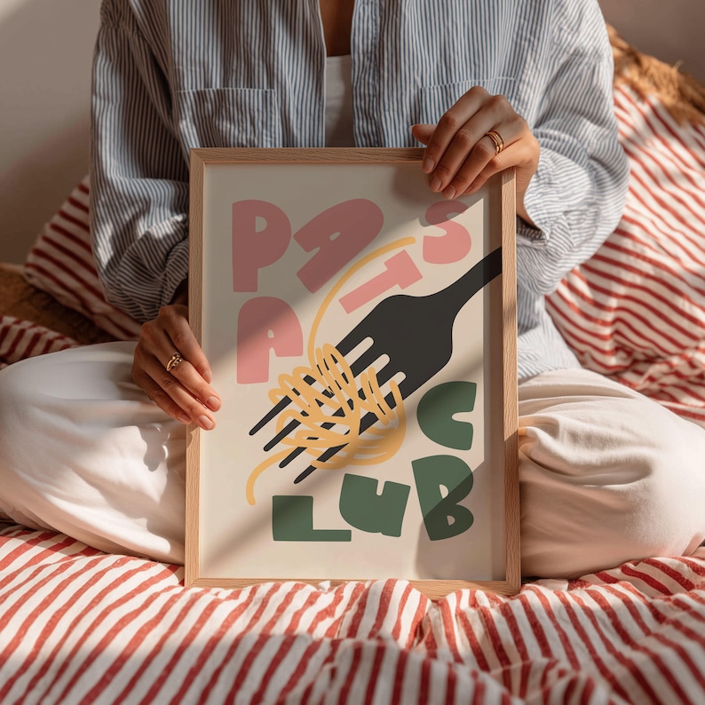 May include: A framed art print with the words "Pasta Club" in pink and green, with a black fork and yellow spaghetti. The print is held by a person sitting on a red and white striped surface.