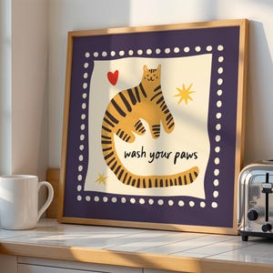K&ouml;nnte beinhalten: Ein quadratisches Wandbild mit Rahmen, das in der cremefarbenen Mitte eine gelbe, tigergestreifte Katze, ein rotes Herz und gelbe Sterne zeigt. Der Text "wash your paws" ist schwarz gedruckt. Der Rand ist dunkelviolett mit wei&szlig;en Punkten.