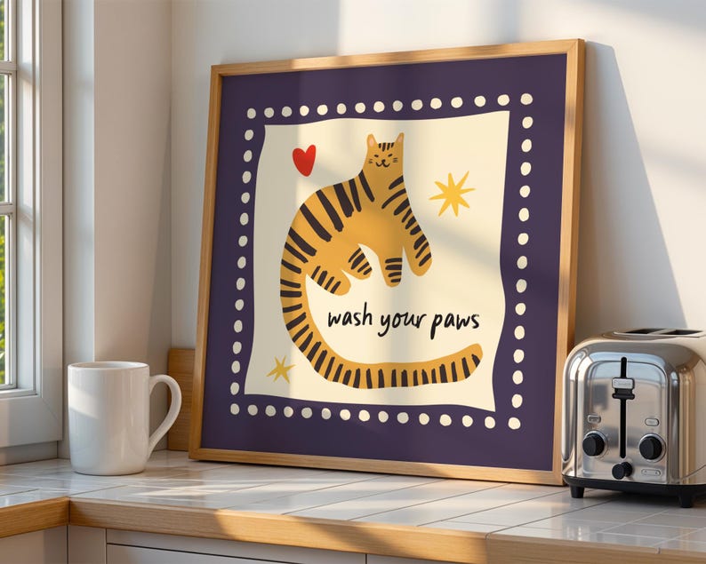 K&ouml;nnte beinhalten: Ein quadratisches Wandbild mit Rahmen, das in der cremefarbenen Mitte eine gelbe, tigergestreifte Katze, ein rotes Herz und gelbe Sterne zeigt. Der Text "wash your paws" ist schwarz gedruckt. Der Rand ist dunkelviolett mit wei&szlig;en Punkten.