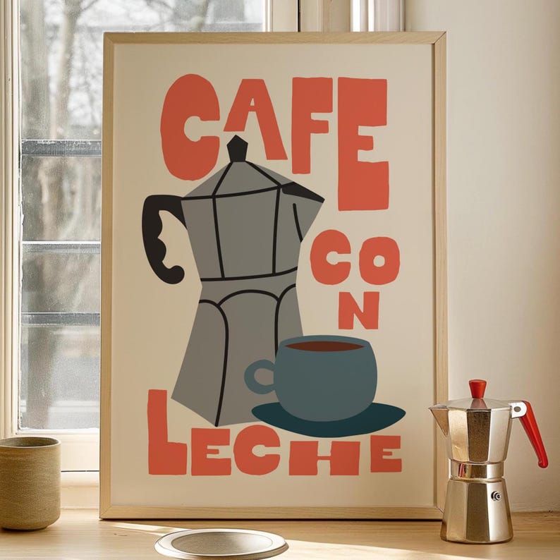 May include: A framed print with the words "CAFE CON LECHE" in orange block letters. The print features a gray coffee maker, a blue cup and saucer, and a cup of coffee. A silver coffee maker with a red handle sits on the right.