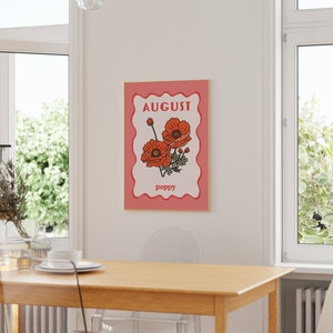 May include: A framed art print featuring a stylized illustration of red poppies. The word "AUGUST" is at the top, and "poppy" is at the bottom. The artwork has a pink border and a gold frame, suitable for wall decor.