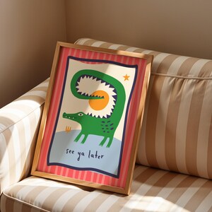 Pu&ograve; includere: Una stampa rettangolare incorniciata raffigurante un coccodrillo verde con la coda curva, un sole giallo e una piccola stella arancione. L'illustrazione include il testo "see ya later" in blu scuro su uno sfondo a righe verticali rosa e rosse.