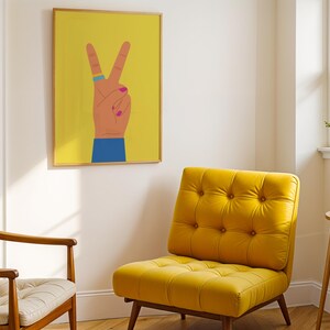 May include: A yellow tufted armchair and a wooden chair with a cream cushion are in a room with a framed print. The print features a hand making a peace sign against a yellow background. The hand has a blue ring and pink nail polish.