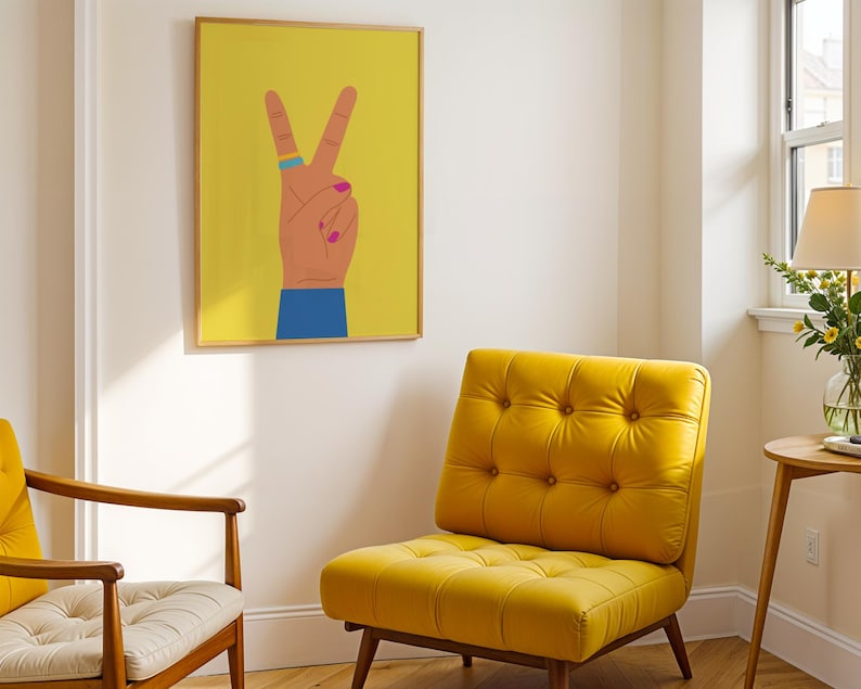 May include: A yellow tufted armchair and a wooden chair with a cream cushion are in a room with a framed print. The print features a hand making a peace sign against a yellow background. The hand has a blue ring and pink nail polish.