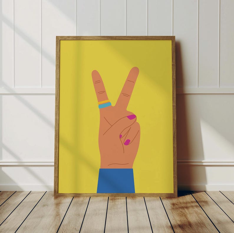 May include: A framed art print featuring a hand making the peace sign against a yellow background. The hand is tan with pink fingernails and a blue and yellow ring. The sleeve is blue.