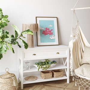 May include: White shelf unit with two shelves and a drawer. The shelf unit has a gold handle on the drawer and is decorated with a framed print that says "You are my sunshine" in pink and red letters on a blue background. There are two woven baskets on the bottom shelf.