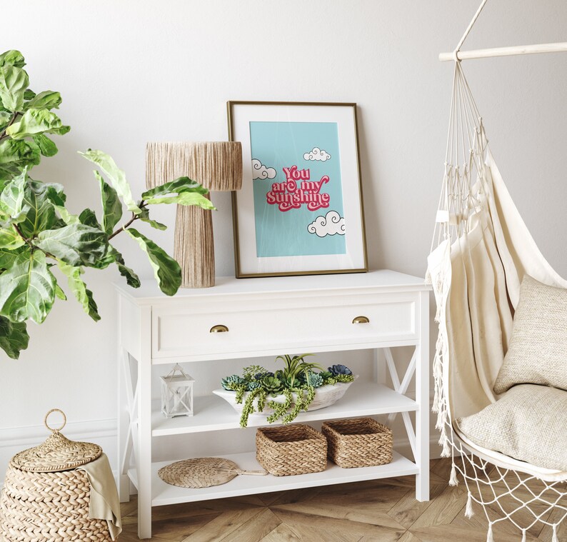 May include: White shelf unit with two shelves and a drawer. The shelf unit has a gold handle on the drawer and is decorated with a framed print that says "You are my sunshine" in pink and red letters on a blue background. There are two woven baskets on the bottom shelf.