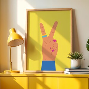 May include: A framed art print featuring a hand making a peace sign against a yellow background. The hand has a blue cuff, a ring with blue and yellow stripes, and pink fingernails. A yellow desk lamp and a small potted plant are also visible.