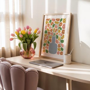 May include: A home office scene features a framed floral print, a vase of tulips, and a laptop. The print has a light wood frame and a colorful floral design. The vase is pink and holds pink and yellow tulips. A speckled ceramic cup and a pink chair are also visible.
