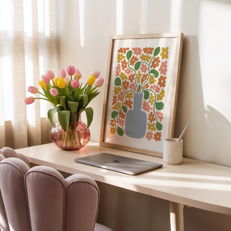 May include: A home office scene features a framed floral print, a vase of tulips, and a laptop. The print has a light wood frame and a colorful floral design. The vase is pink and holds pink and yellow tulips. A speckled ceramic cup and a pink chair are also visible.