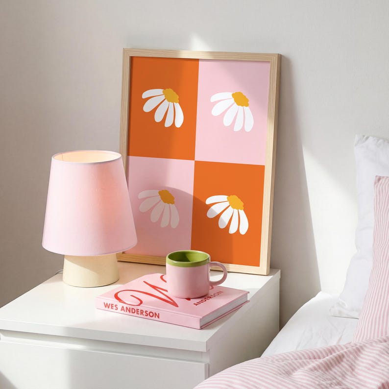 May include: A framed art print with a daisy design in orange and pink squares. A pink lamp, a pink mug with a green rim, and a book with the title "WES ANDERSON" are on a white nightstand.