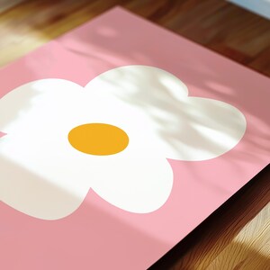 May include: A pink rectangular mat featuring a large white flower with a yellow center. The flower design is centered on the mat. The mat is placed on a wooden surface, with shadows cast across the top.