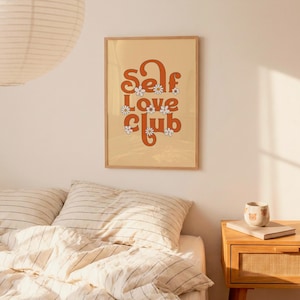 May include: A rectangular wall art print in a light wooden frame featuring the text Self Love Club in bold orange retro-style lettering. The design is decorated with small white daisy flowers with yellow centres against a light beige background.