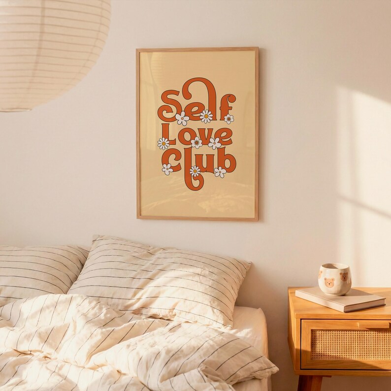 May include: A rectangular wall art print in a light wooden frame featuring the text Self Love Club in bold orange retro-style lettering. The design is decorated with small white daisy flowers with yellow centres against a light beige background.