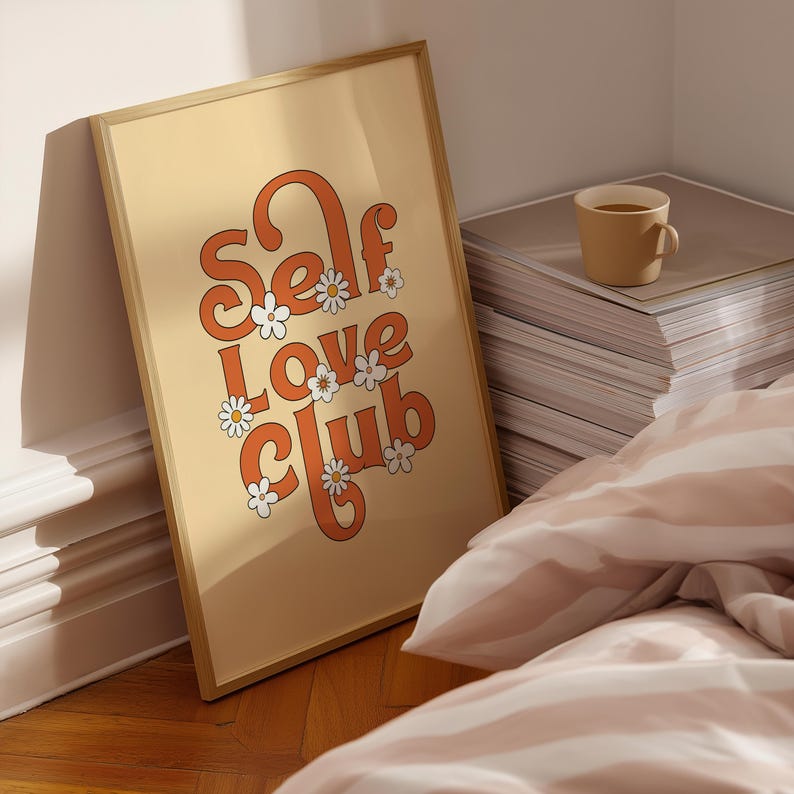 May include: A rectangular framed wall art print featuring a light beige background with the orange text "Self Love Club" in a retro-inspired font. The design is decorated with small white daisy flowers with yellow centres scattered throughout the letters.
