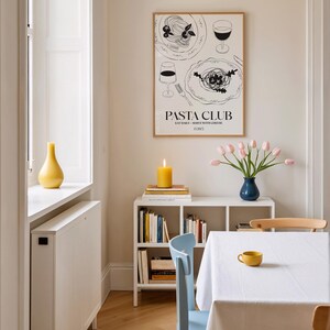 K&ouml;nnte beinhalten: Esszimmer-Szene mit einem gerahmten "Pasta Club"-Poster mit Abbildungen von Pastagerichten und Weingl&auml;sern. Eine brennende gelbe Kerze steht auf B&uuml;chern. Eine blaue Vase h&auml;lt rosa Tulpen, und eine gelbe Tasse ruht auf einer wei&szlig;en Tischdecke.