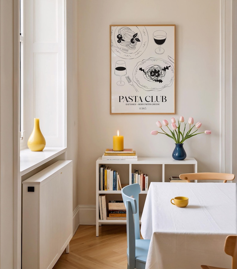 K&ouml;nnte beinhalten: Esszimmer-Szene mit einem gerahmten "Pasta Club"-Poster mit Abbildungen von Pastagerichten und Weingl&auml;sern. Eine brennende gelbe Kerze steht auf B&uuml;chern. Eine blaue Vase h&auml;lt rosa Tulpen, und eine gelbe Tasse ruht auf einer wei&szlig;en Tischdecke.