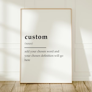 Op de afbeelding: Een rechthoekige wandkunstprint met een lay-out in de stijl van een woordenboekdefinitie. De tekst luidt custom (noun) add your chosen word and your chosen definition will go here. De print is ingelijst in een dunne houten lijst.