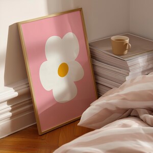 May include: A framed art print featuring a large white flower with a yellow center on a pink background. The artwork is in a gold frame and leans against a white wall. A beige mug sits on a stack of papers.