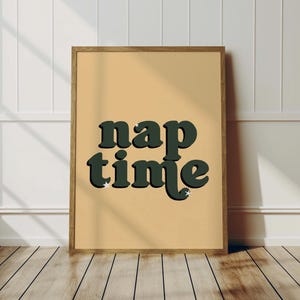 May include: A framed print featuring the words "nap time" in dark green, retro lettering with a white outline and star accents. The background is a light tan, and the frame is a warm brown colour.