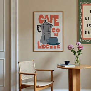 May include: A framed art print with a coffee theme. The print features the words "CAFE CON LECHE" in coral-colored block letters, along with a gray coffee maker and a blue cup of coffee. The print is in a wooden frame.