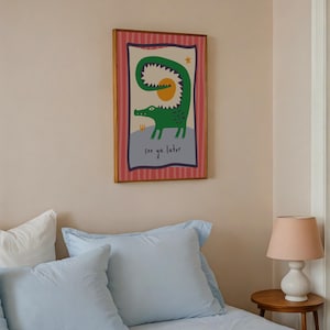 Pu&ograve; includere: Una stampa artistica da parete rettangolare incorniciata raffigurante un coccodrillo verde con la coda curva, un sole giallo e una piccola stella su sfondo chiaro. Il bordo &egrave; rosa a righe verticali e il testo see ya later &egrave; in basso.