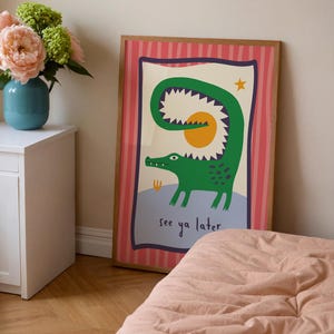 Pu&ograve; includere: Una stampa da parete rettangolare incorniciata raffigurante un coccodrillo verde con la coda curva che forma un cerchio attorno a un sole giallo. L'illustrazione include una piccola stella gialla e il testo see ya later in blu scuro su sfondo azzurro.