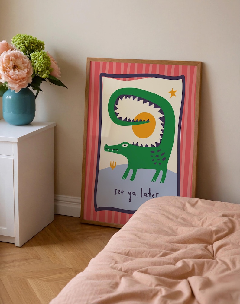 Pu&ograve; includere: Una stampa da parete rettangolare incorniciata raffigurante un coccodrillo verde con la coda curva che forma un cerchio attorno a un sole giallo. L'illustrazione include una piccola stella gialla e il testo see ya later in blu scuro su sfondo azzurro.