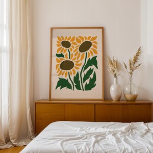 May include: A framed art print featuring a stylized illustration of three sunflowers in shades of yellow, brown, and green. The artwork is displayed above a wooden cabinet, with vases of dried plants. The room has a neutral color palette.