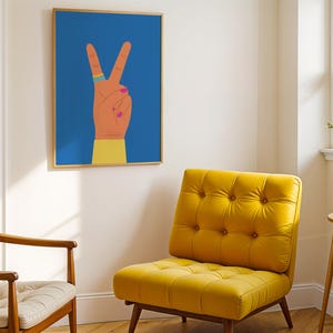 May include: A framed art print featuring a hand making the peace sign against a blue background. The hand is depicted with orange skin, pink nail polish, and a ring. A yellow upholstered chair and a wooden chair are in the foreground.