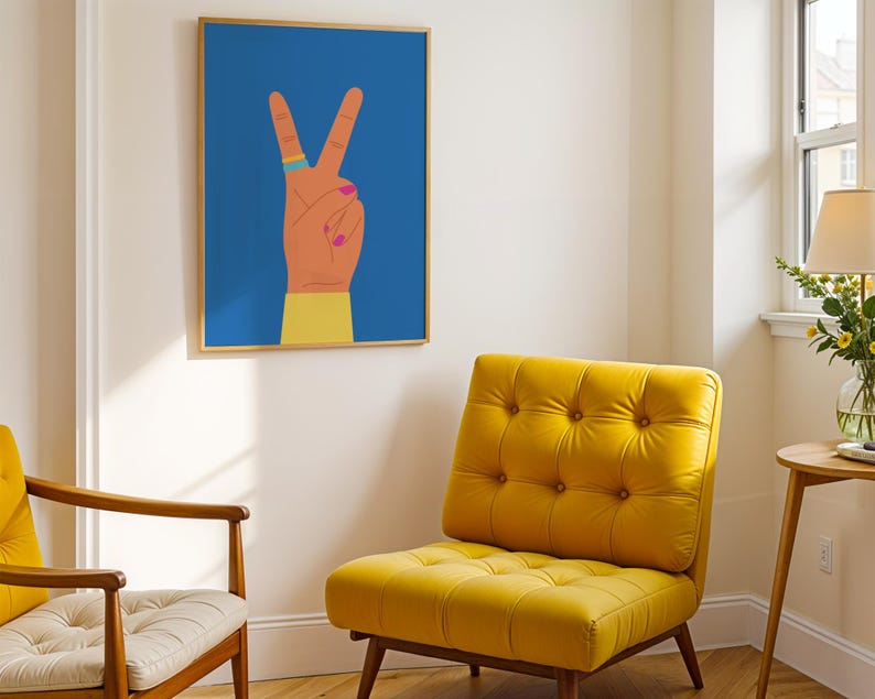 May include: A framed art print featuring a hand making the peace sign against a blue background. The hand is depicted with orange skin, pink nail polish, and a ring. A yellow upholstered chair and a wooden chair are in the foreground.