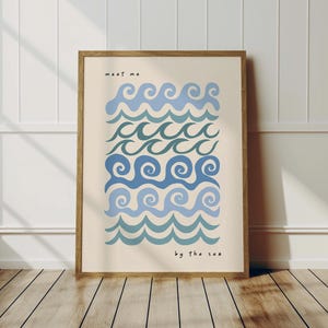 May include: A framed art print featuring stylized blue wave patterns in varying shades against a cream background. The words "meet me" and "by the sea" are printed in black. The frame is a light brown color.