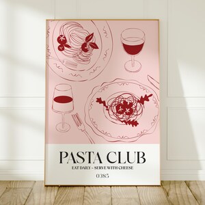 K&ouml;nnte beinhalten: Ein gerahmtes Poster mit rosa Hintergrund zeigt Strichzeichnungen von Pastagerichten, Weingl&auml;sern und einer Gabel. Der Text "PASTA CLUB" steht unten, darunter "EAT DAILY - SERVE WITH CHEESE". Das Poster steht auf einem Holzboden.