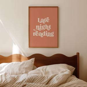 May include: A framed art print with the words "Late night reading" in white script on a salmon-colored background. The wooden frame hangs above a bed with striped bedding and a dark wooden headboard.