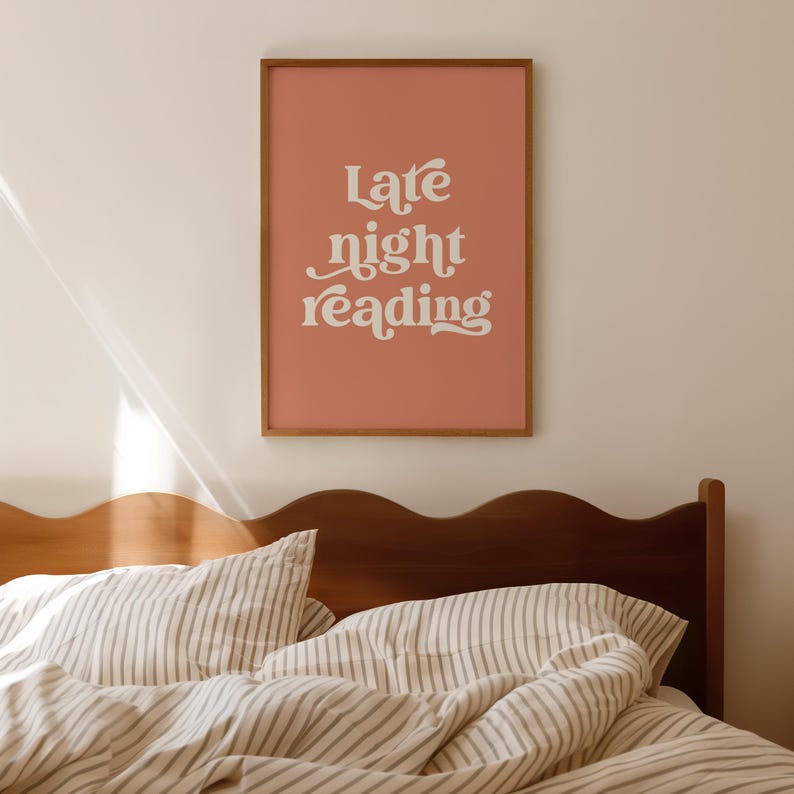 May include: A framed art print with the words "Late night reading" in white script on a salmon-colored background. The wooden frame hangs above a bed with striped bedding and a dark wooden headboard.