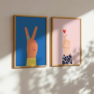 May include: Two framed art prints. One features a hand making a peace sign against a blue background. The other shows a hand forming a heart shape with a red heart on a pink background. Both have wooden frames.