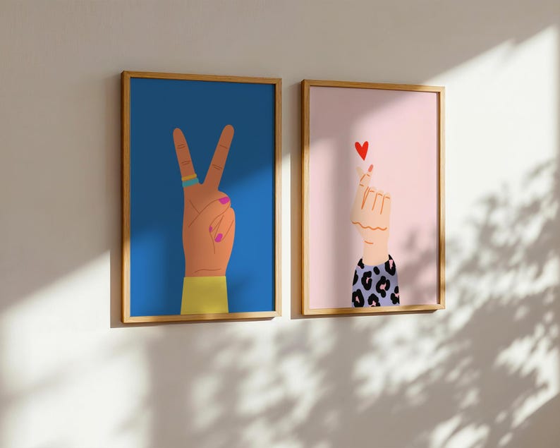 May include: Two framed art prints. One features a hand making a peace sign against a blue background. The other shows a hand forming a heart shape with a red heart on a pink background. Both have wooden frames.