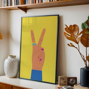 May include: A framed art print featuring a hand making the peace sign against a yellow background. The hand has a blue ring, pink nail polish, and a blue sleeve. The artwork is displayed on a white surface with other decorative items.
