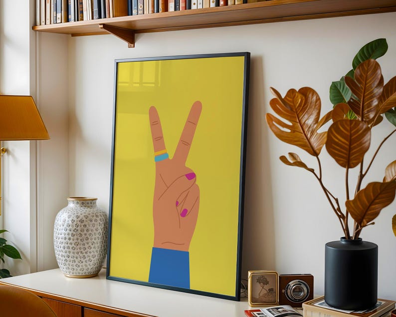 May include: A framed art print featuring a hand making the peace sign against a yellow background. The hand has a blue ring, pink nail polish, and a blue sleeve. The artwork is displayed on a white surface with other decorative items.