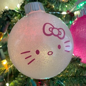 Op de afbeelding: Een glinsterende witte kerstbal met een paars Hello Kitty gezicht. De bal hangt aan een groene kerstboom, versierd met kleurrijke lichtjes.