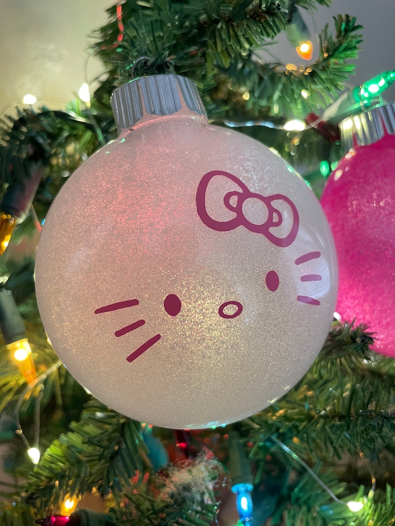 Op de afbeelding: Een glinsterende witte kerstbal met een paars Hello Kitty gezicht. De bal hangt aan een groene kerstboom, versierd met kleurrijke lichtjes.