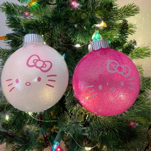 Op de afbeelding: Twee glinsterende kerstballen, &eacute;&eacute;n witte en &eacute;&eacute;n roze, elk versierd met een roze Hello Kitty gezichtsontwerp. De ballen hangen aan een groene kerstboom met kleurrijke lichtjes, wat een feestelijke sc&egrave;ne cre&euml;ert.