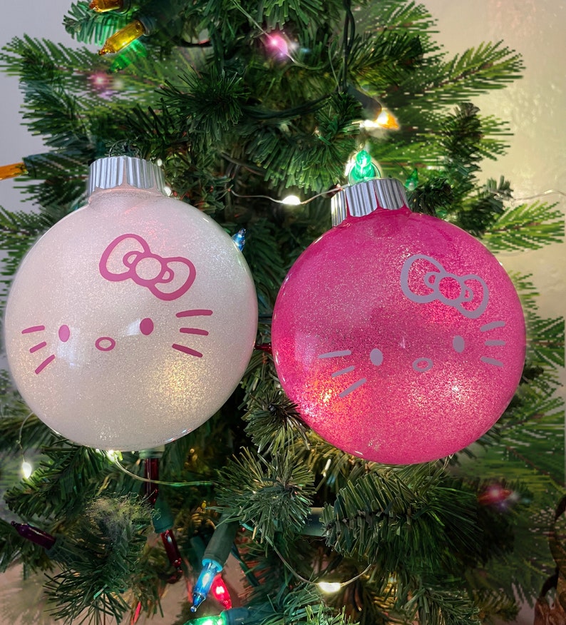 Op de afbeelding: Twee glinsterende kerstballen, &eacute;&eacute;n witte en &eacute;&eacute;n roze, elk versierd met een roze Hello Kitty gezichtsontwerp. De ballen hangen aan een groene kerstboom met kleurrijke lichtjes, wat een feestelijke sc&egrave;ne cre&euml;ert.