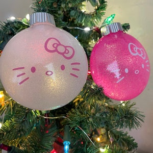 Op de afbeelding: Twee glinsterende kerstballen met Hello Kitty-gezichten hangen aan een kerstboom. E&eacute;n ornament is wit, de andere roze. Beide hebben zilveren tops en zijn versierd met de iconische strik, snorharen en gezichtskenmerken.