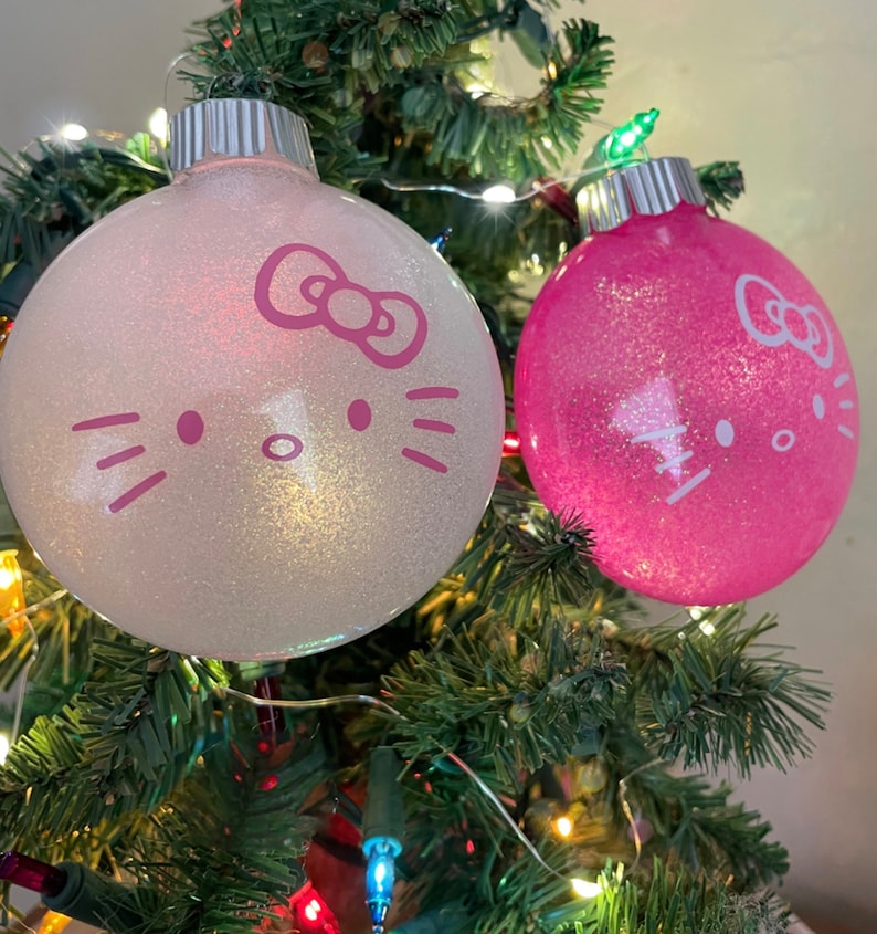 Op de afbeelding: Twee glinsterende kerstballen met Hello Kitty-gezichten hangen aan een kerstboom. E&eacute;n ornament is wit, de andere roze. Beide hebben zilveren tops en zijn versierd met de iconische strik, snorharen en gezichtskenmerken.