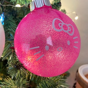 Op de afbeelding: Een roze glitter kerstbal met een zilveren dop. De ornament heeft een witte afbeelding van een kattengezicht met een strik. De bal hangt aan een groene kerstboom.